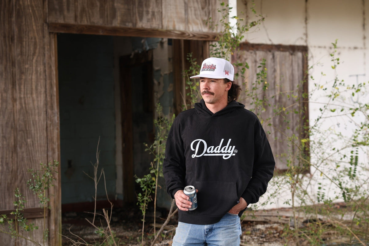A man with a mustache wearing a white "Boobs Make Me Happy" trucker hat and a black hoodie holding a drink outdoors.