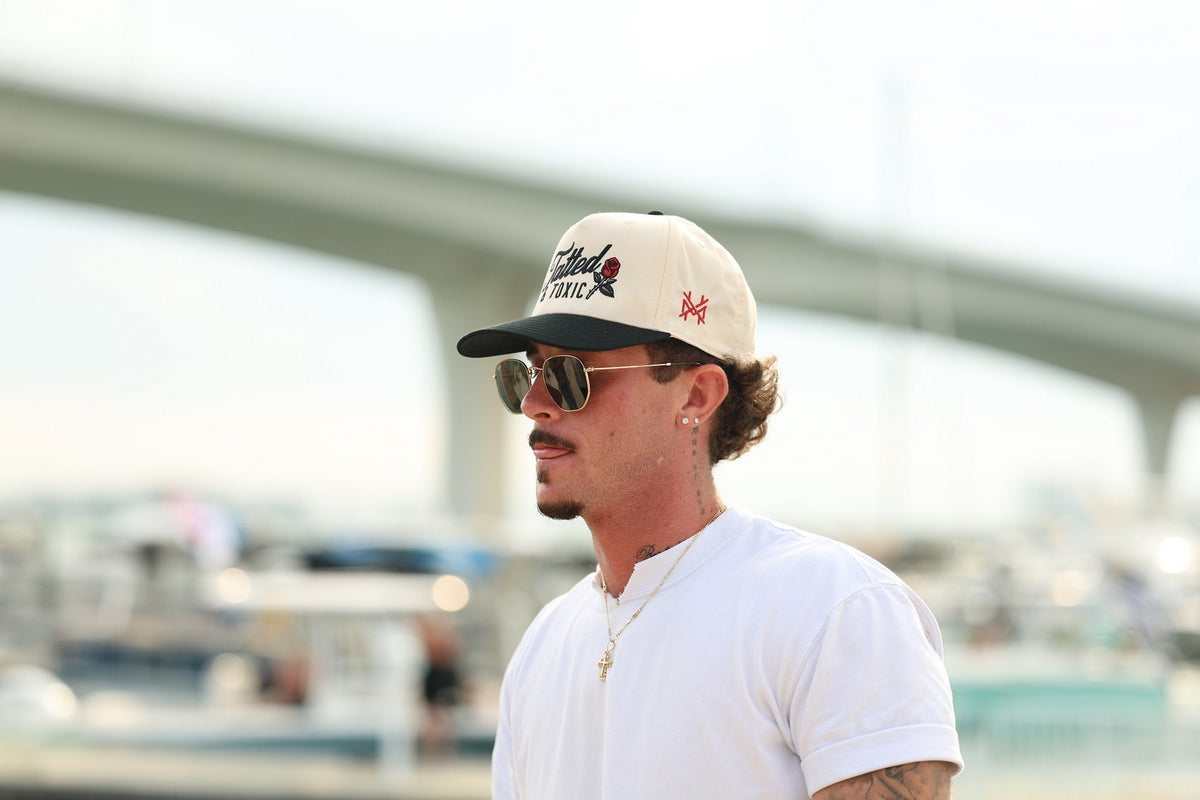 A man wearing a white t-shirt, sunglasses, and a two-tone "Tatted & Toxic" snapback hat outdoors near a bridge and marina.