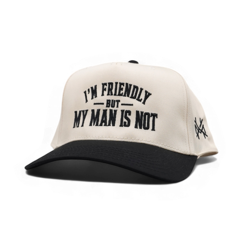 An off-white and black trucker-style cap with the text 'I'M FRIENDLY BUT MY MAN IS NOT' embroidered on the front, and a small black logo on the side, shown on a plain white background.